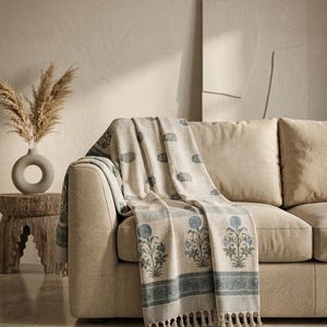 May include: A beige throw blanket featuring a repeating blue floral block print pattern, finished with a decorative border and fringe along the bottom edge. The fabric displays a light, neutral base color with muted blue botanical motifs.