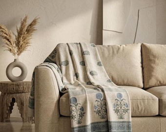 Puede incluir: Una manta beige con un patrón floral azul repetido en estampado de bloques, terminada con un borde decorativo y flecos en el borde inferior. La tela muestra un color base neutro y claro con motivos botánicos en azul apagado.
