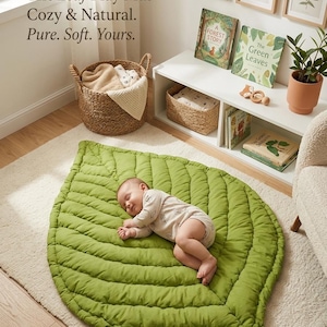 Puede incluir: Una alfombra de juego verde con forma de hoja y detalles de costuras acolchadas. El texto dice: The Leaf Play Mat - Cozy & Natural. Pure. Soft. Yours. Un beb&eacute; duerme sobre la alfombra, que est&aacute; colocada sobre una alfombra clara.