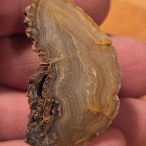 May include: A polished, semi-translucent agate stone with layered bands of tan, cream, and brown. The rough, textured edge contrasts with the smooth, polished surface. The stone is held in a hand, showcasing its natural beauty.