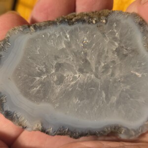 May include: A polished, irregularly shaped agate stone slice. The stone features a translucent, light gray center with concentric bands of white and blue-gray. The outer edge is a rough, dark brown. The stone is held in a hand.