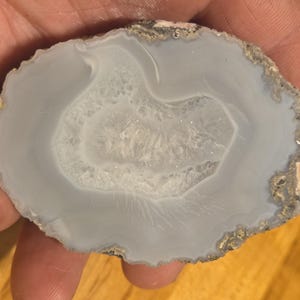 May include: A polished, oval-shaped agate stone slice. The stone features concentric bands of light blue and white, with a crystalline center. The outer edge has a rough, textured appearance with hints of brown and gray.