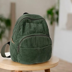 May include: A dark green corduroy backpack featuring a rounded main compartment with a zip closure, a smaller front zipped pocket, and a side pocket with a tan trim. The bag has dark green straps and a textured, ribbed fabric surface.