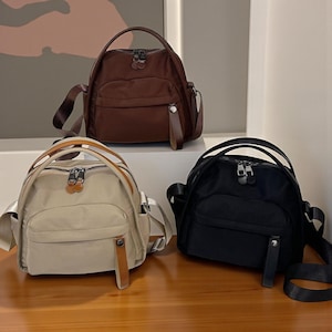 Peut inclure: Trois petits sacs à dos arrondis, marron, beige et noir. Chaque sac est doté d'une poignée supérieure, d'une poche avant et d'une longue bandoulière réglable. Les sacs ont des fermetures éclair argentées et des accents marron simili cuir.
