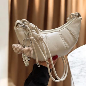 May include: Cream-colored crescent-shaped handbag with a zipper closure and stitched details. The bag features a knotted handle and a decorative keychain with a bow and pearl accents. The bag is held by a person.