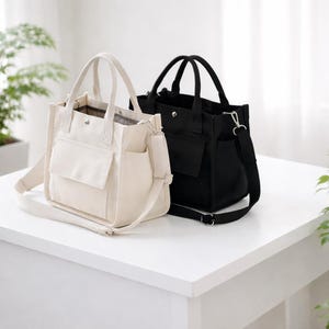 Könnte beinhalten: Zwei Canvas-Tragetaschen, eine cremefarbene und eine schwarze, stehen nebeneinander. Jede Tasche hat einen oberen Griff, einen abnehmbaren Schultergurt und eine Außentasche. Die cremefarbene Tasche hat eine Vordertasche und eine Seitentasche. Die schwarze Tasche hat eine Vordertasche.