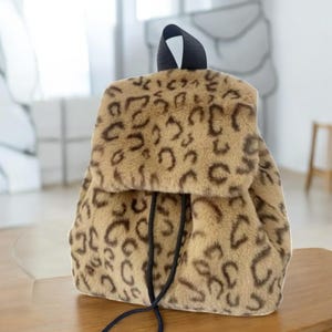May include: A faux fur backpack featuring a tan and brown leopard print pattern. The bag has a fold-over flap closure, a black top handle, and black drawstring cords that hang down the front of the main compartment for secure fastening.