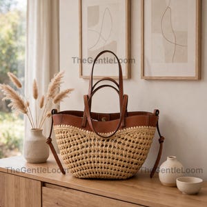 May include: A tote bag featuring a woven tan straw base and a smooth brown leather upper section with two matching leather handles. The text TheGentleLoom is visible on the bag and in the background. The bag has a wide, open-top design.