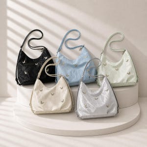 May include: Five shoulder bags in black, light blue, cream, silver, and pale green are displayed. Each bag features a butterfly motif and a long strap. The bags are arranged on a white, circular platform.