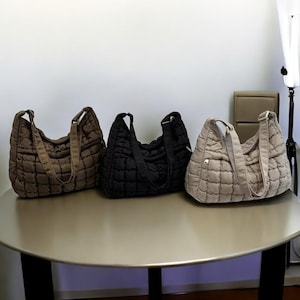 Könnte beinhalten: Drei gesteppte Schultertaschen in Braun, Schwarz und Beige stehen auf einem runden Tisch. Jede Tasche hat eine rechteckige Form mit einem Steppmuster und einem langen Riemen. Die Taschen sind aus weichem, gepolstertem Material.