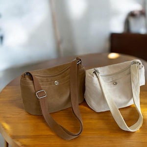 Può includere: Sono mostrate due borse a tracolla in tela. Una è di colore marrone chiaro, l'altra è bianco sporco. Entrambe le borse hanno una forma rettangolare con chiusura a zip e tracolle regolabili. Le borse sono su un tavolo di legno.