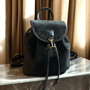 Könnte beinhalten: Schwarzer Rucksack aus Wildleder mit gebogenem Überschlag und oberem Griff. Die Tasche hat einen Kordelzugverschluss und goldfarbene Hardware. Verstellbare Schultergurte und ein kleiner Griff sind sichtbar.
