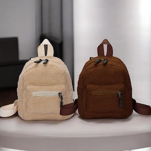Könnte beinhalten: Zwei kleine, flauschige Rucksäcke. Einer ist hellbeige mit einem cremefarbenen Griff und Riemen, der andere ist ein sattes Braun mit passendem braunen Griff und Riemen. Beide haben eine Vordertasche mit Reißverschluss und schwarze Reißverschlüsse.