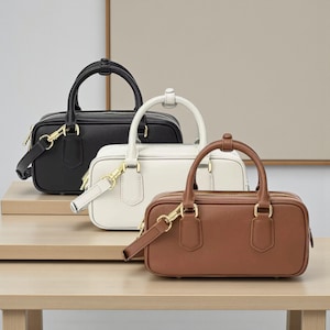 May include: Three small, rectangular handbags in black, white, and brown. Each bag has a top handle and a detachable shoulder strap. The bags feature gold-coloured hardware, including zips and clasps. They are displayed on a light-coloured wooden surface.