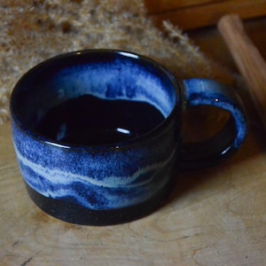 May include: A ceramic mug with a dark blue interior and a blue and white wave pattern on the exterior. The mug has a black base and a rounded handle. A wooden spoon rests beside the mug.