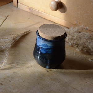 May include: A small, handmade ceramic jar with a cork lid. The jar is dark blue with lighter blue streaks, and the cork lid is a light brown. The jar is sitting on a wooden surface, with dried plants nearby. The jar is approximately 10 cm tall.