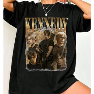 May include: Black t-shirt featuring a graphic design with the text "KENNEDY" in gold lettering. The design includes multiple images of a person in various poses. The shirt is made of a soft material.