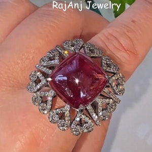 May include: A large, ornate ring featuring a square, deep red gemstone at its center. The gemstone is surrounded by a detailed, silver-toned setting with smaller, sparkling stones. The ring is from RajAnj Jewelry.