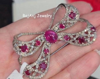 May include: A silver-toned bow brooch with a central oval ruby and smaller ruby accents. The brooch is encrusted with small, clear gemstones. The text "RajAnj Jewelry" is visible. A tag with text is attached.