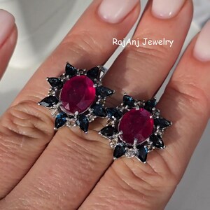 Puede incluir: Pendientes de plata con un rubí ovalado central, rodeado de piedras de acento azul oscuro y transparente. Los pendientes tienen un diseño de estrella. El texto "RajanJ Jewelry" es visible.