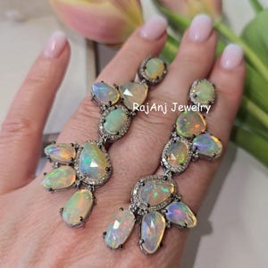 May include: A pair of silver earrings featuring multiple opal gemstones in various shapes, surrounded by small clear stones. The earrings have a cluster design with the text "RajAnj Jewelry".