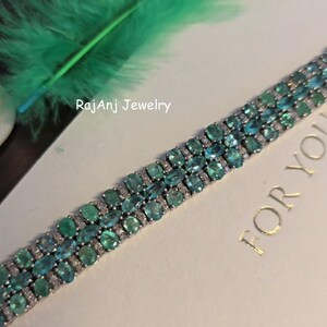 May include: A close-up of a silver bracelet with rows of oval-cut, teal-colored gemstones. The bracelet is set with smaller, clear stones between the larger gems. The text "RajAnj Jewelry" is visible in the image.