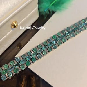 May include: A three-row bracelet featuring oval-cut emerald and teal gemstones, interspersed with small, clear stones. The bracelet is set in a silver-colored metal and is displayed on a white surface. The text "RajanJ Jewelry" is visible.