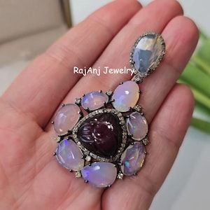 May include: A handcrafted pendant featuring a central, dark red gemstone surrounded by opals and small diamonds. The pendant has a silver-tone setting and the text "RajanJ Jewelry".