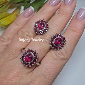 Op de afbeelding: Drie ringen met een ovale robijn in het midden, omringd door kleinere robijnen en witte edelstenen. De ringen zijn gezet op een zilveren band. De tekst "RajAnj Jewelry" is zichtbaar.