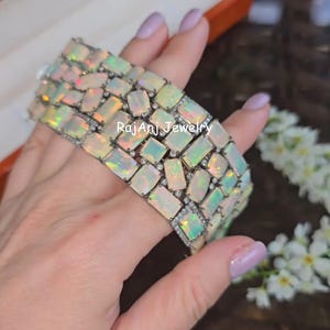 Può includere: Un bracciale largo con un design a mosaico di opali rettangolari. Gli opali mostrano una gamma di colori, tra cui bianco, giallo e verde. Piccole pietre trasparenti sono intercalate. Il testo "Rajan Jewelry" è visibile.