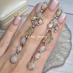 Può includere: Orecchini pendenti lunghi con un gruppo di opali di varie forme. Gli opali mostrano colori iridescenti e sono incastonati in un metallo argentato con piccole pietre chiare. Orecchini di RajAnj Jewelry.
