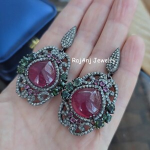 May include: A pair of elaborate earrings with large, teardrop-shaped, ruby-red gemstones. The earrings have a dark metal setting, with smaller green and pink stones, and a delicate, openwork design. The top section is a smaller, elongated shape, also encrusted with small stones.