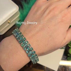 May include: A silver bracelet with rows of oval-cut, teal-colored gemstones and small, clear accent stones. The bracelet is on a wrist, with the text "RajAnj Jewelry" visible in the background.