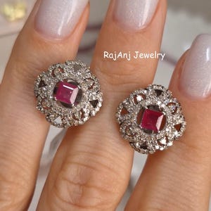 May include: A pair of silver-toned stud earrings. Each earring features a square, deep red gemstone at the center, surrounded by a halo of smaller, clear gemstones. The earrings have an openwork design around the central stones.