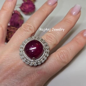 May include: A large, ornate ring featuring a vibrant, oval-shaped, deep red gemstone. The gemstone is surrounded by a halo of smaller, sparkling stones set in a silver-toned metal. The ring is displayed on a hand, with the text "RajAnj Jewelry" visible.