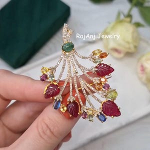 Puede incluir: Broche dorado con diseño de abanico, adornado con una gran gema verde y varias gemas coloridas. El broche presenta gemas rojas, amarillas y azules, y está incrustado con pequeñas piedras transparentes. El texto "Rajanj Jewelry" es visible.