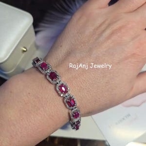 Peut inclure: Un bracelet argenté orné de pierres précieuses rectangulaires rouge foncé, chacune entourée de petites pierres claires. Le bracelet est porté au poignet. Le texte "RajanJ Jewelry" est visible.
