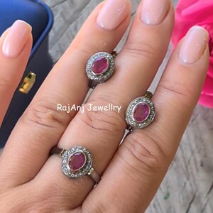May include: Three silver-toned rings, each featuring an oval-cut red gemstone surrounded by a halo of small clear stones. The rings are displayed on fingers, and the text RajAnj Jewellry is visible in the center of the image.
