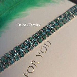 May include: A silver bracelet with rows of oval-cut teal gemstones and small clear stones. The bracelet is on a white surface with the text "FOR YOU" and "RajanJ Jewelry". A green feather is in the background.
