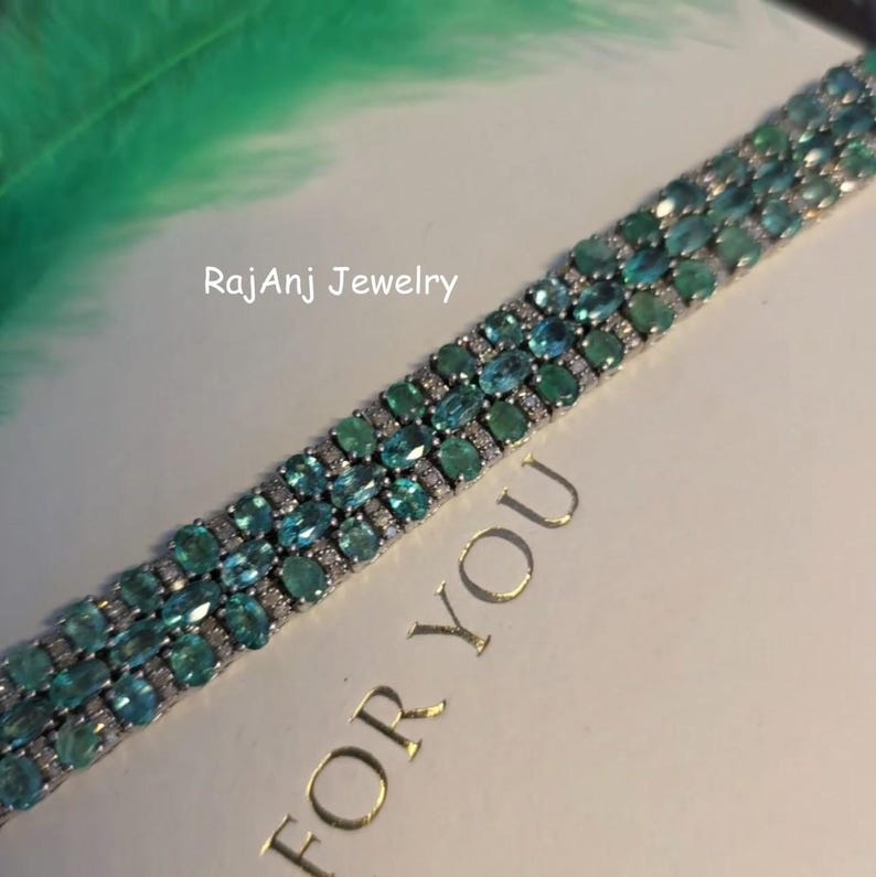 May include: A silver bracelet with rows of oval-cut teal gemstones and small clear stones. The bracelet is on a white surface with the text "FOR YOU" and "RajanJ Jewelry". A green feather is in the background.