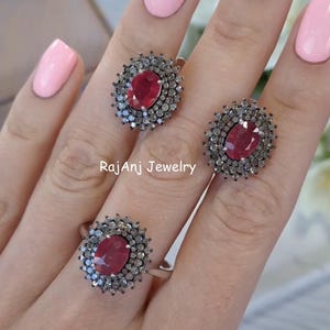 May include: A set of three rings featuring oval-cut, deep red gemstones surrounded by a halo of smaller, silver-toned stones. The rings are displayed on a hand with pink nail polish. The text "RajAnj Jewelry" is visible.