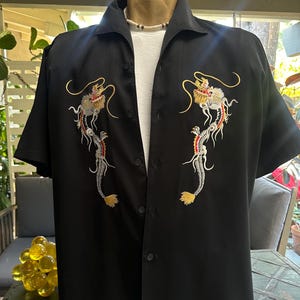 May include: A black short-sleeved button-down shirt featuring symmetrical embroidery of two dragons on the chest. The dragons are stitched in gold, red, white, and black thread. The shirt has a classic collar and is worn over a plain white undershirt.