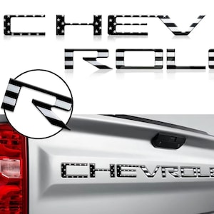 May include: A set of individual black and white letters spelling CHEVROLET, featuring a pattern of stars on the first half and horizontal stripes on the second half. The letters are shown applied to the silver tailgate of a pickup truck.