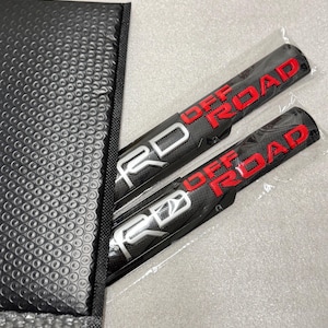 May include: Two rectangular automotive emblems featuring a black carbon fibre texture. Each emblem displays the silver TRD logo on the left and the red text OFF ROAD on the right. The emblems are encased in clear plastic protective sleeves.