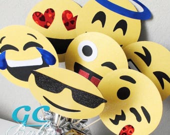 May include: A set of seven yellow emoji-themed party decorations. The emojis include a laughing emoji with tears, a winking emoji with sunglasses, a tongue-sticking-out emoji, a heart-eyed emoji, a smiling emoji with a halo, a winking emoji, and a kissing emoji.