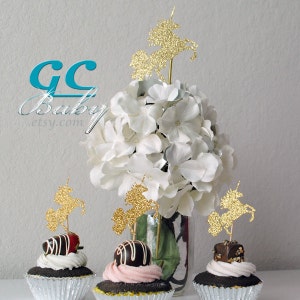 Op de afbeelding: Gouden glitter-eenhoorn cupcake-toppers op cupcakes met chocolade frosting en witte hortensia's in een glazen vaas. De toppers zijn perfect voor een eenhoorn-themafeest.