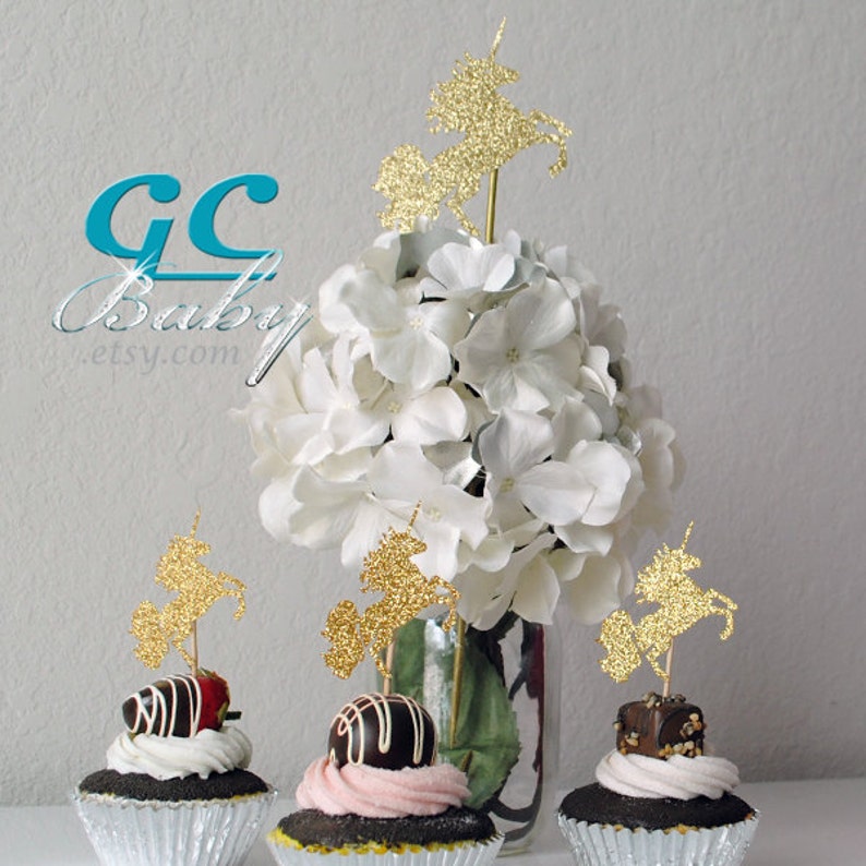 Op de afbeelding: Gouden glitter-eenhoorn cupcake-toppers op cupcakes met chocolade frosting en witte hortensia's in een glazen vaas. De toppers zijn perfect voor een eenhoorn-themafeest.