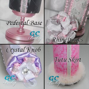 May include: A pink pedestal base with a crystal knob and a white tutu skirt with rhinestones. The base is decorated with a pink and white bow and the skirt is decorated with a pink and white damask pattern.