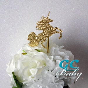 Op de afbeelding: Gouden glitter unicorn taarttopper. De unicorn staat op zijn achterpoten met zijn hoorn omhoog. De topper staat op een wit bloemstuk.