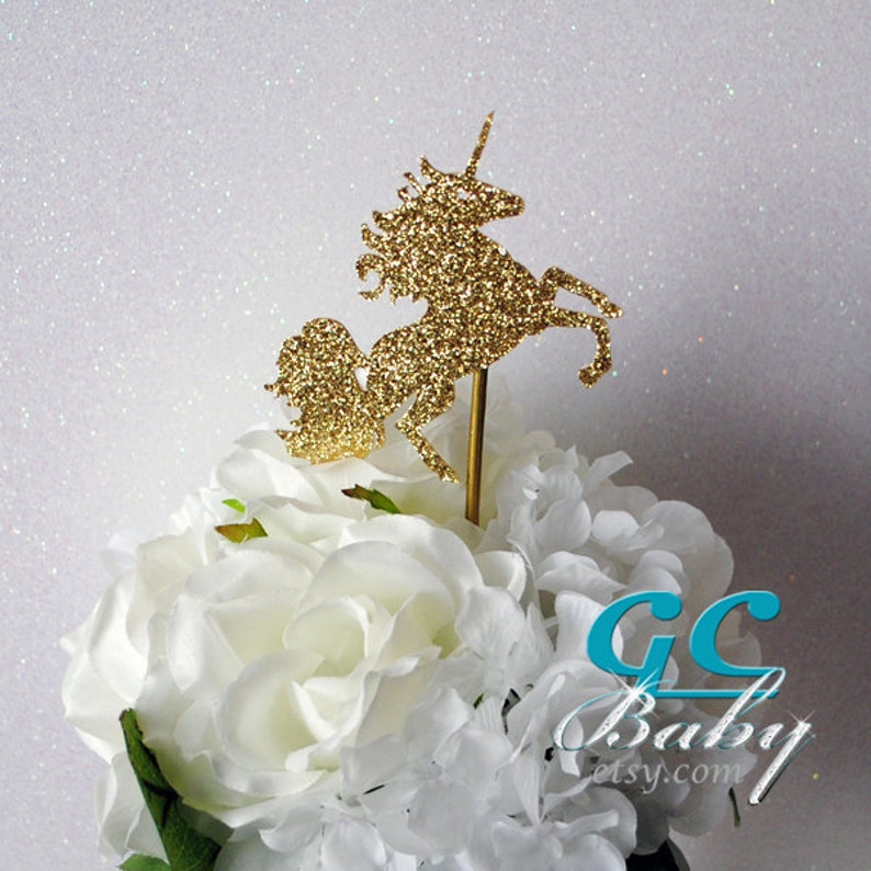Op de afbeelding: Gouden glitter unicorn taarttopper. De unicorn staat op zijn achterpoten met zijn hoorn omhoog. De topper staat op een wit bloemstuk.